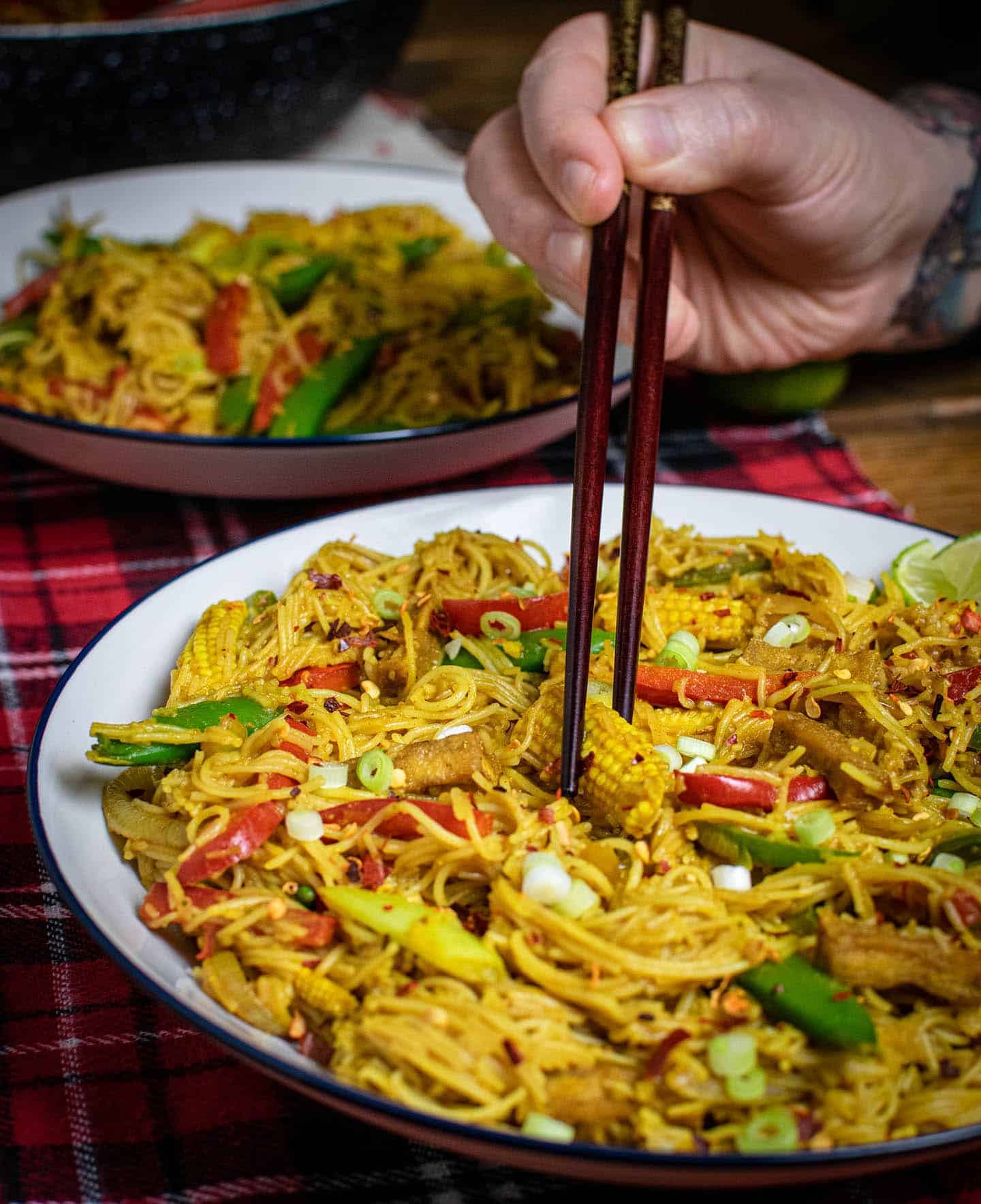 Veg Singapore noodles on a white plate with a pair of hands holding chopsticks, about to pick up some baby corn to eat,