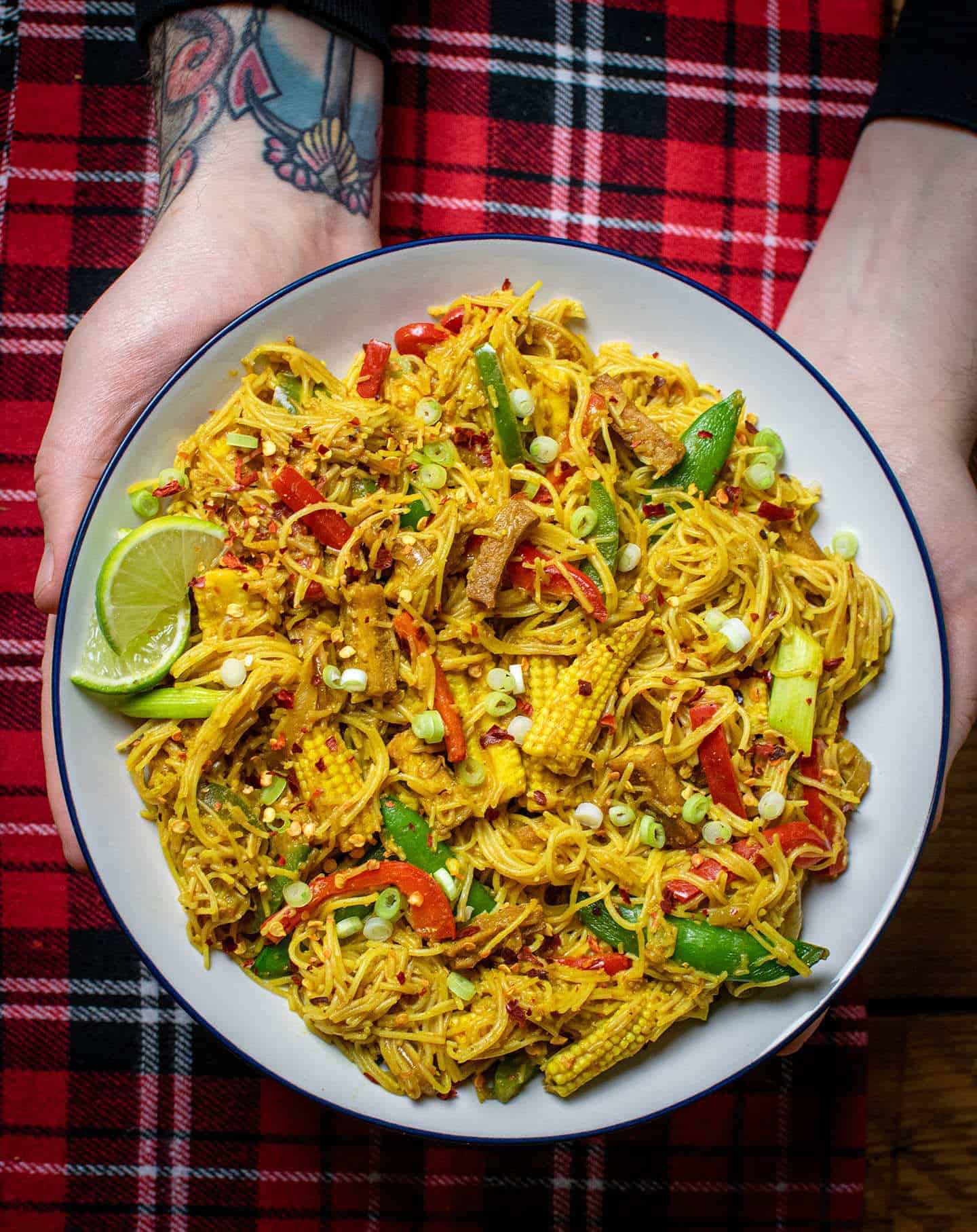 A pair of hands with a tattoo on the left wrist holding a plate of veg Singapore Noodles, with two slices of limes as a garnish on the left of the plate.