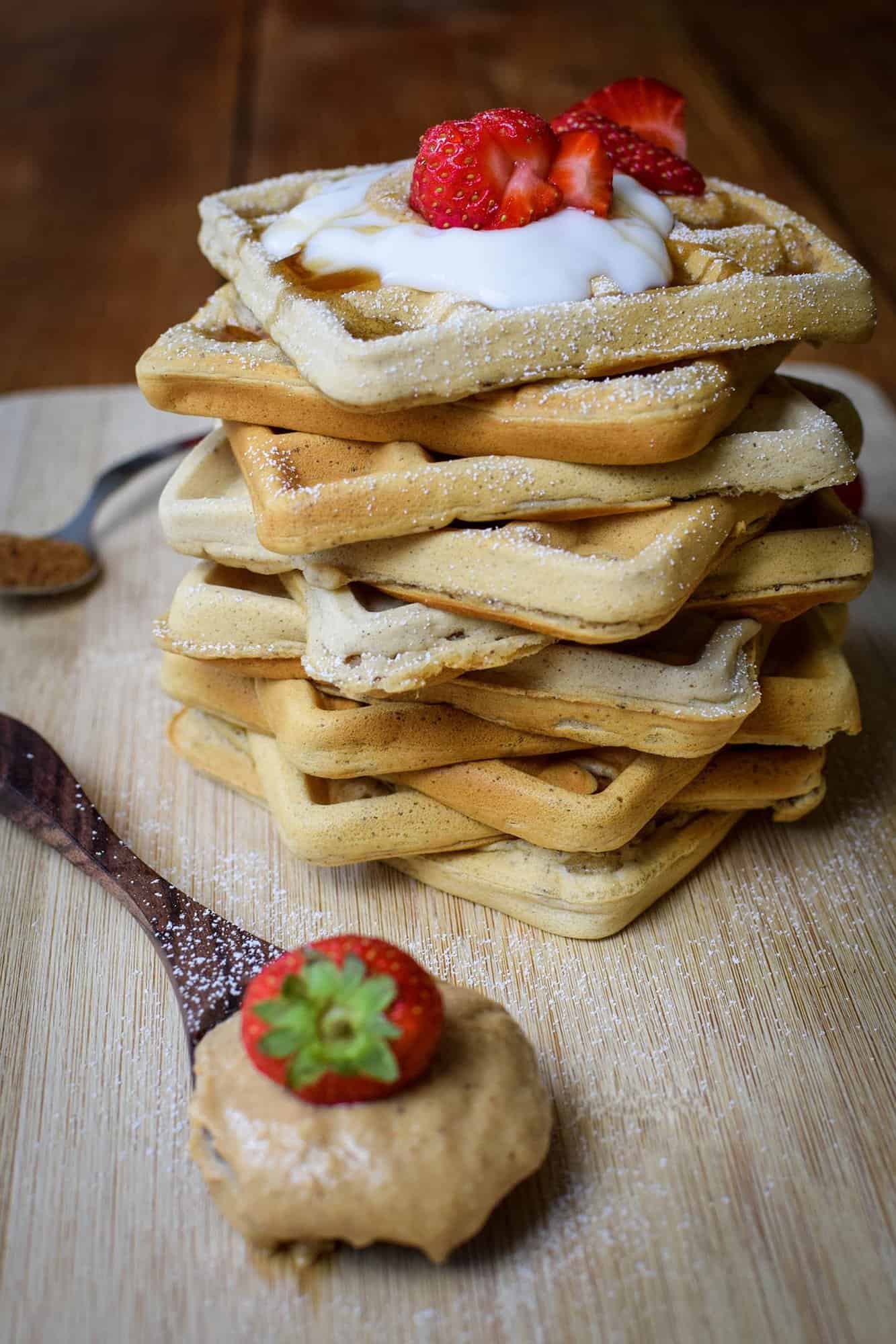 simple vegan waffles stacked up, with a wooden spoon in the foreground loaded with peanut butter and a strawberry