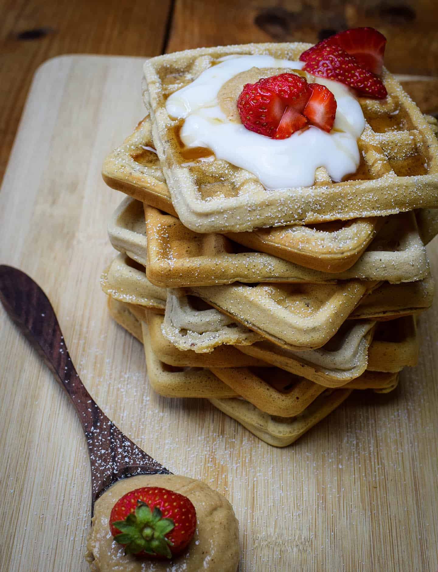 simple vegan waffles stacked up. Birds eye view of strawberries, coconut yoghurt and peanut butter.There's a spoon in the background with sugar on it.