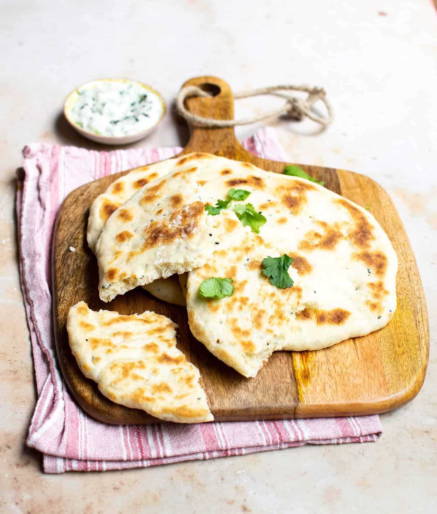 Naan bread on a wooden chopping board with a pink cloth underneath and raita in the background