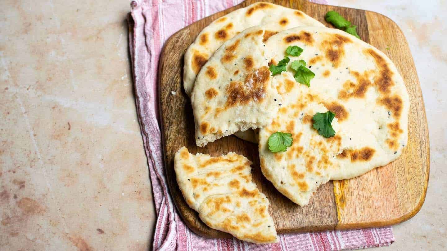 Landscape image of naan bread on a wooden board with coriander on top, the bread is ripped on top with whole ones underneath