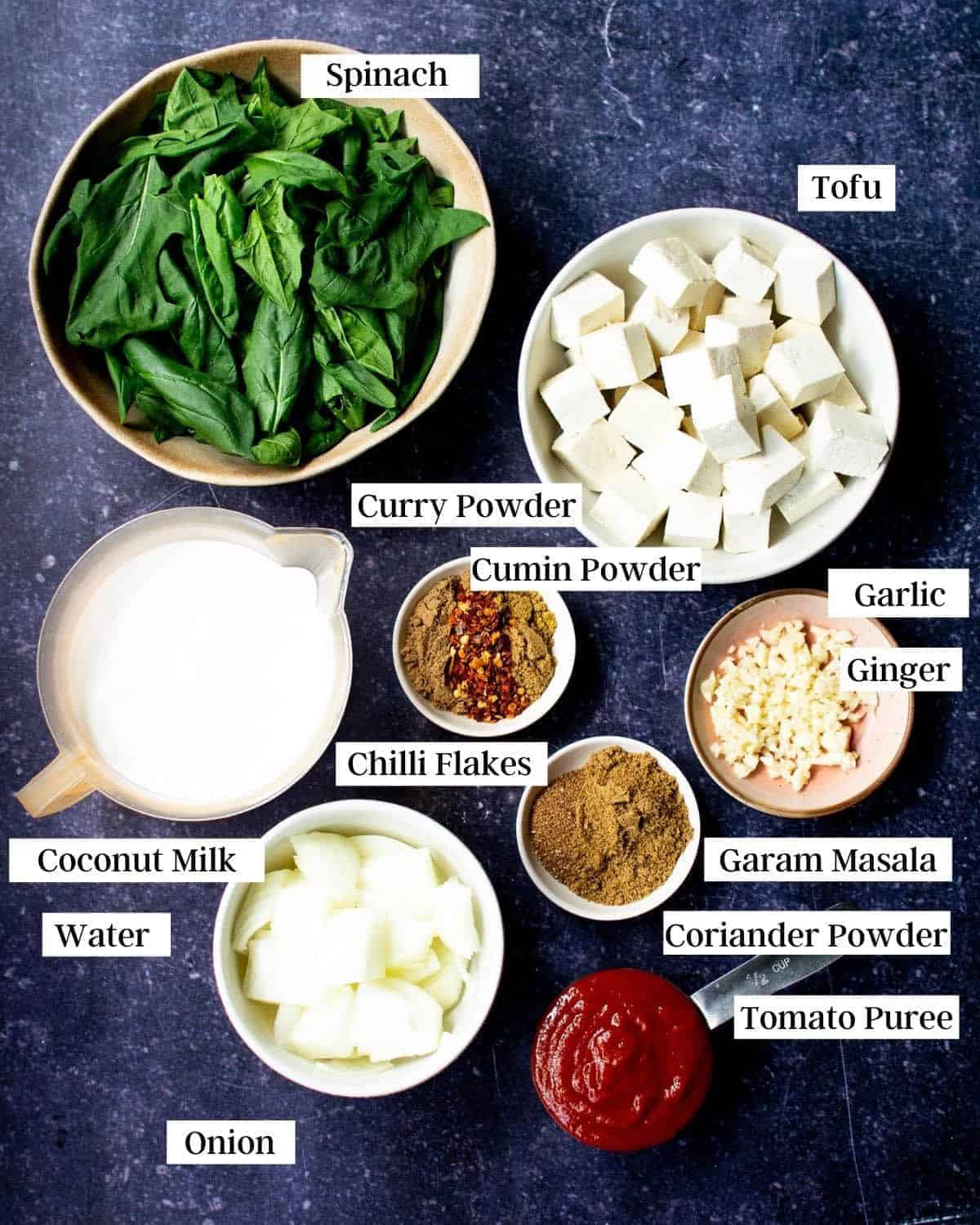 Ingredients for vegan palak tofu laid out on a blue background with labels by each item.