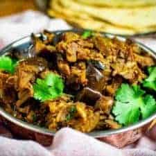 Brinjal bhaji in a classic balti dish surrounded by a pink tea towel. There's coriander on top of the dish and chapatis in the background