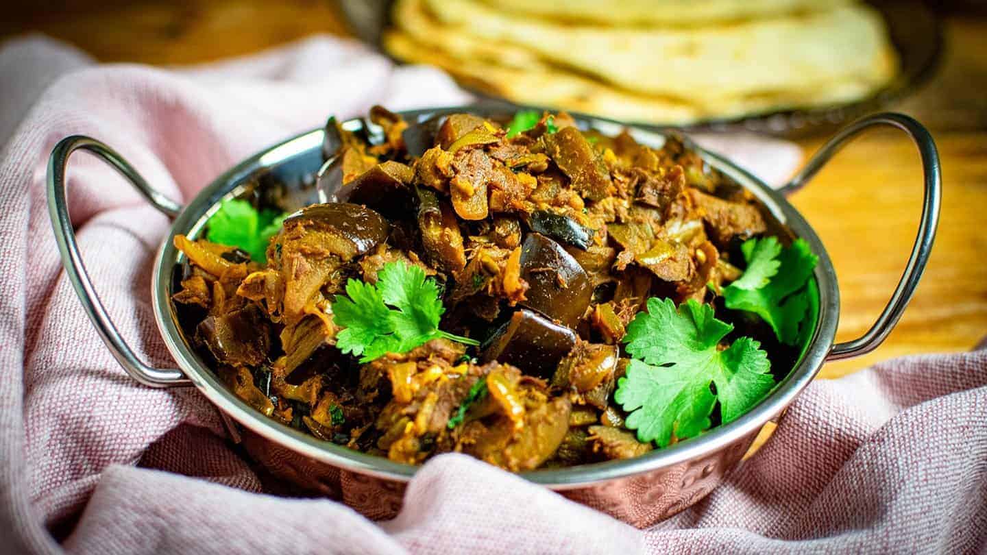 Brinjal bhaji in a classic balti dish surrounded by a pink tea towel. There's coriander on top of the dish and chapatis in the background