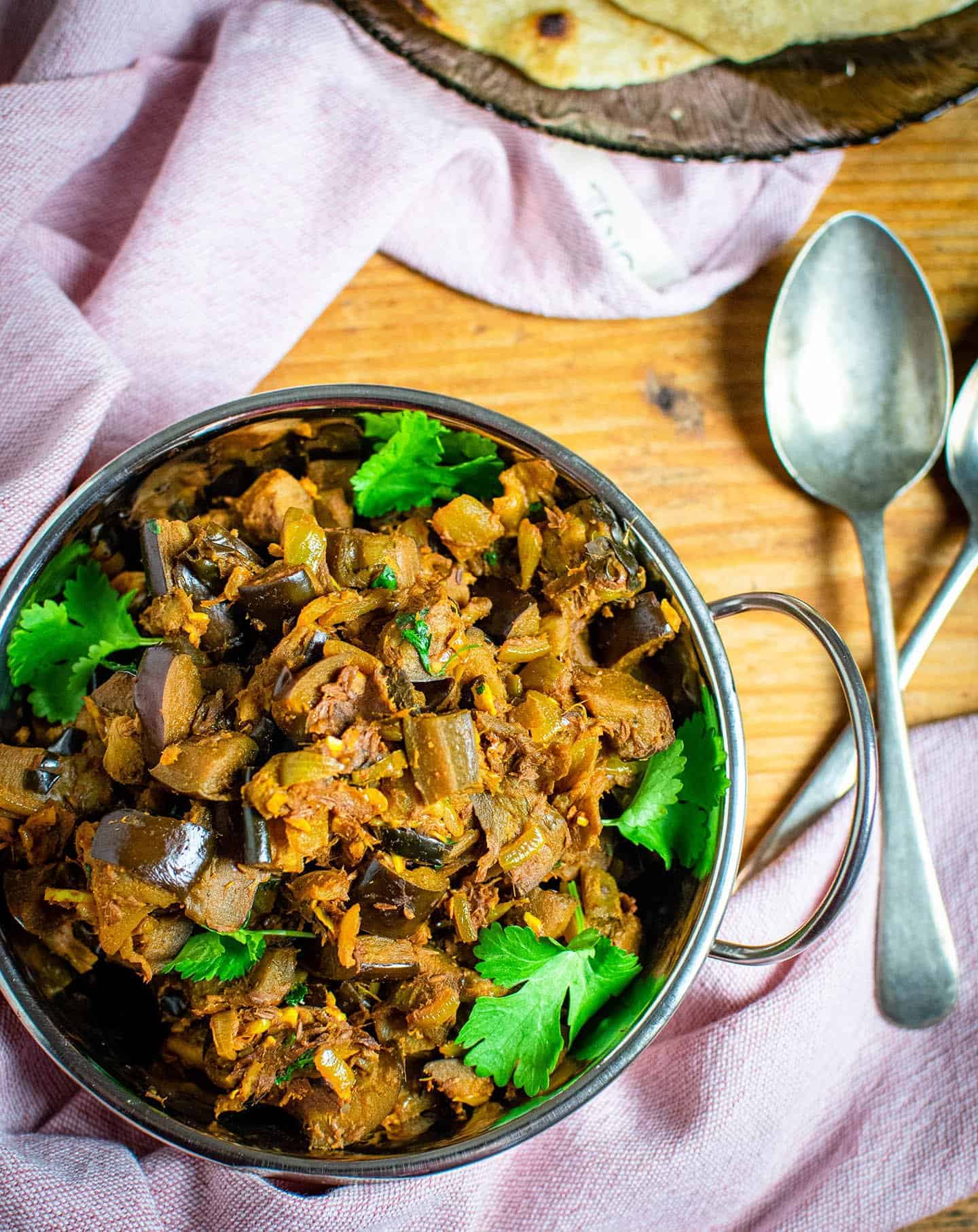 Flat lay image of the aubergine bhaji, spoons are visible to the right, two handles are crossed over each other. A pink tea towel can also be seen.