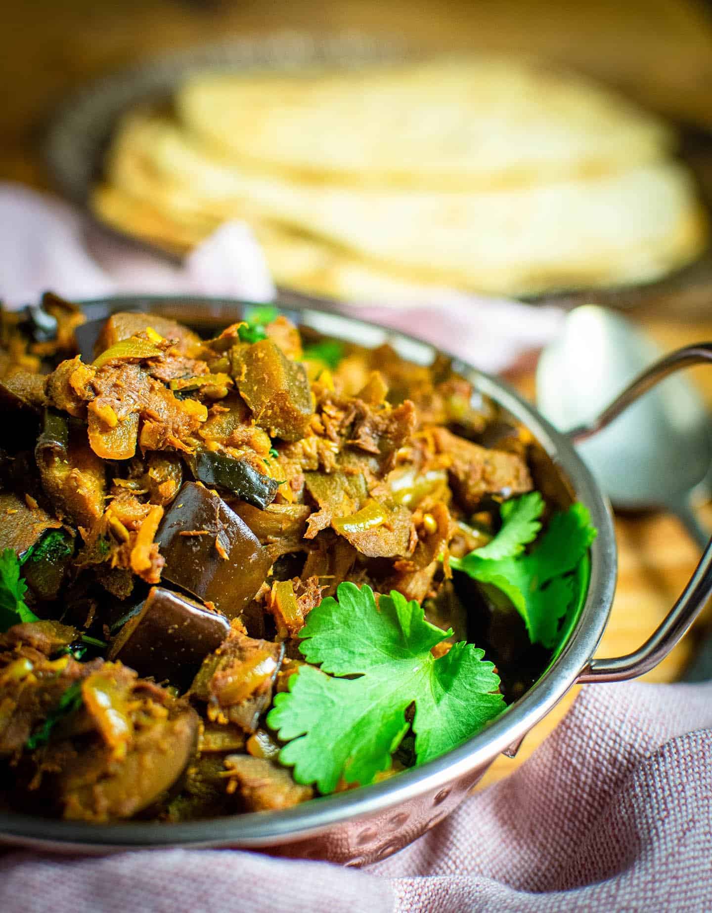 Brinjal bhaji in a dish with fresh corainder. Spoons and a plate of chaptis are in the background.