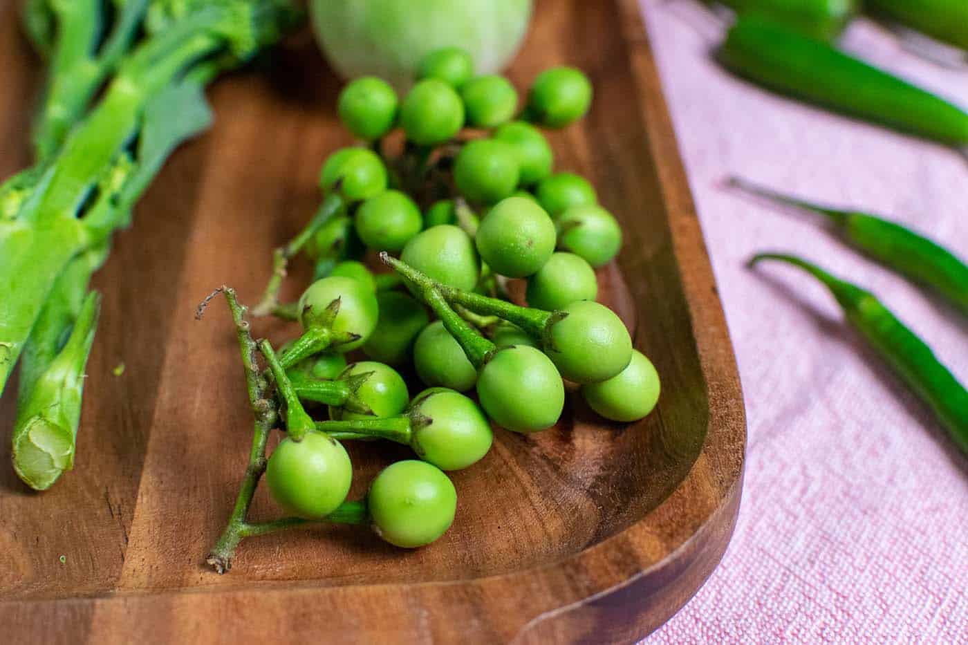 Pea eggplants on a wooden chopping board with broccoli and Thai eggplants in the background