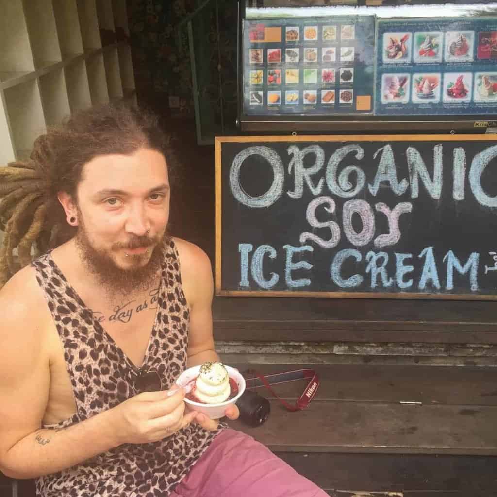 Dan sitting in front of an organic soy ice cream parlour in Ubud Bali, holding ice cream and a sign in the background says 'Organic Soy Ice Cream'