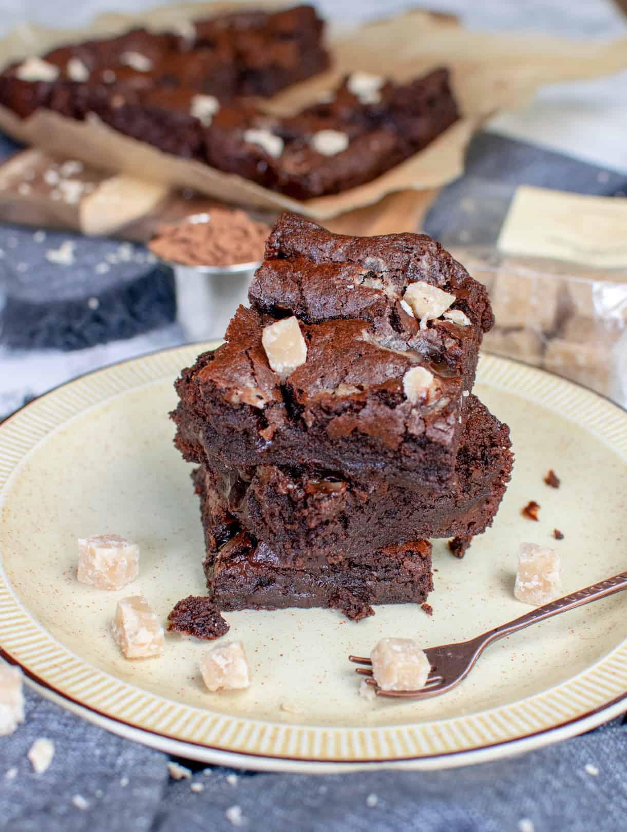 3 pieces of fudge brownie stacked on top of each other topped with vegan fudge, which is also scattered around the yellow plate