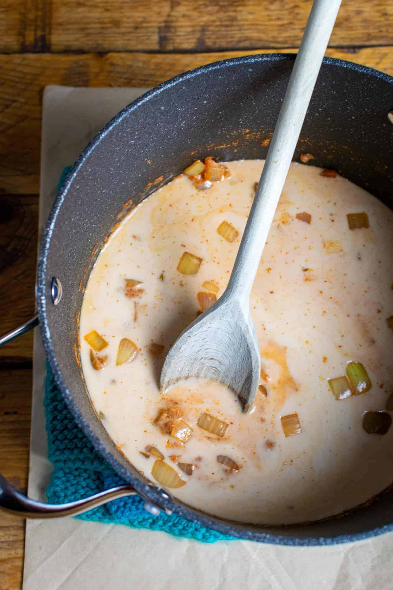 Vegan pumpkin soup in a large soup pan, showing the coconut milk, onion, curry paste in the pan with a wooden spoon