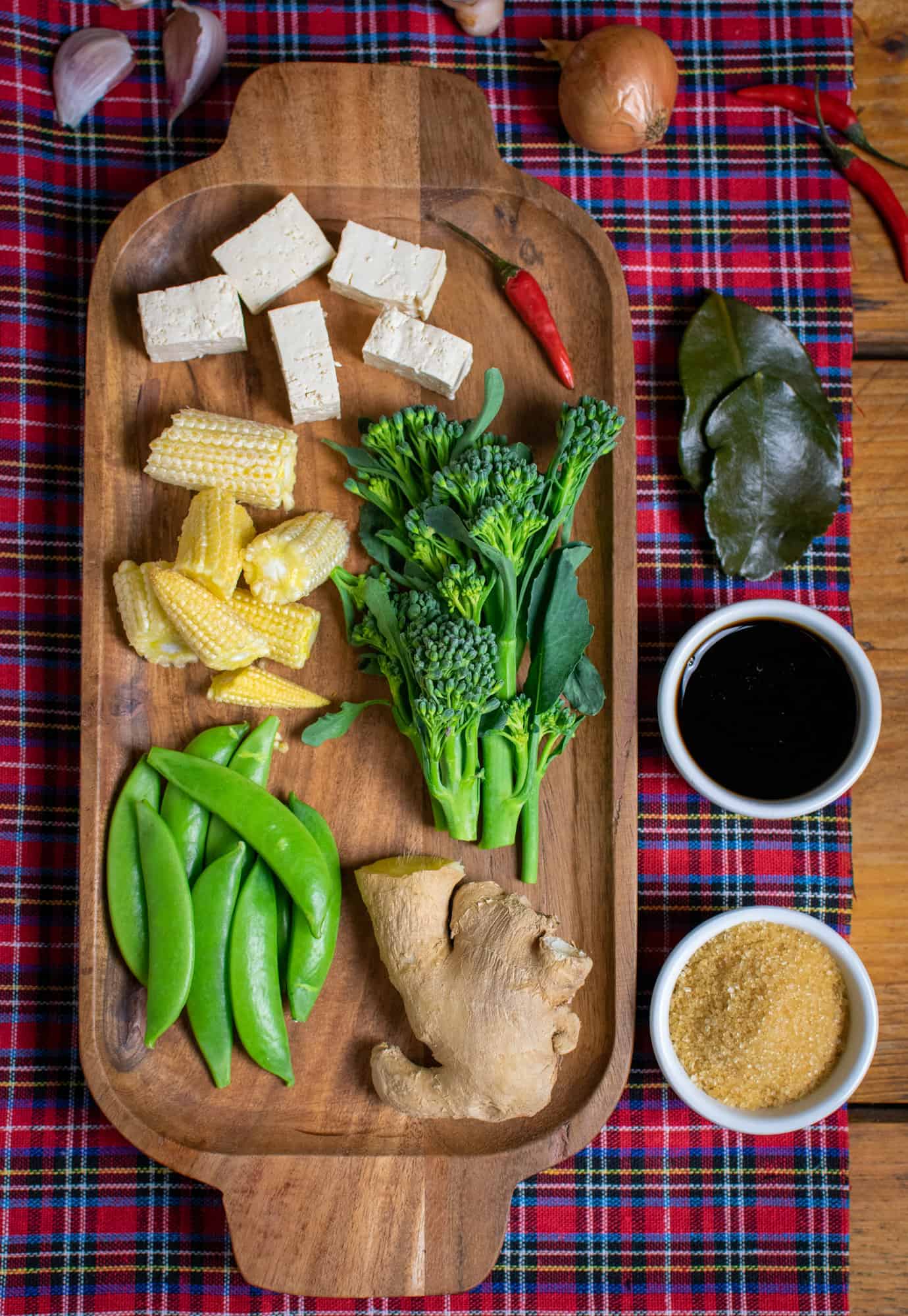 Various vegetables like tenderstem broccoli, sugar snap peas and baby corn on a wooden tray with tofu, ginger a chilli and kaffir lime leaves as well as soy sauce and brown sugar