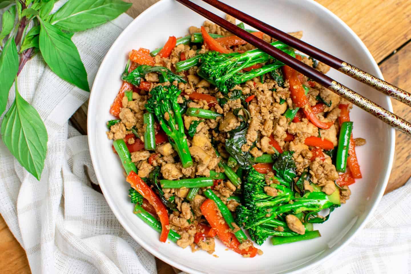 A landscape picture of a white bowl filled with Thai basil tofu stir-fry. Crumbled tofu and fresh vegetables are all visible, including tenderstem broccoli, green beans, red pepper and wilted holy basil. Next to the bowl is a linen cloth and a bunch of fresh holy basil.