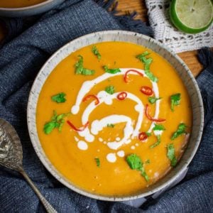 Vegan pumpkin soup in a bowl, with oat cream on top and chillies and coriander. Set on top of a blue towel with a lime in the background and spoon to the left.