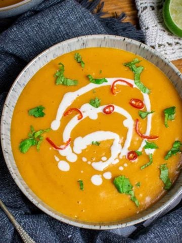 Vegan pumpkin soup in a bowl, with oat cream on top and chillies and coriander. Set on top of a blue towel with a lime in the background and spoon to the left.