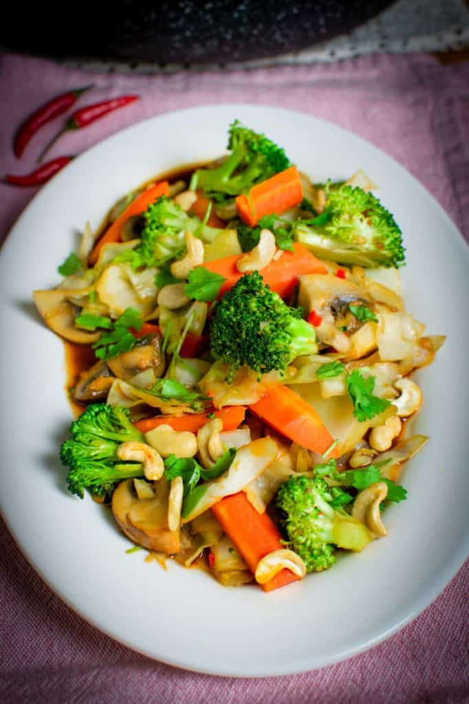 Thai vegetable stir fry on a oval white plate sat on a pink towel, with a pan in the background