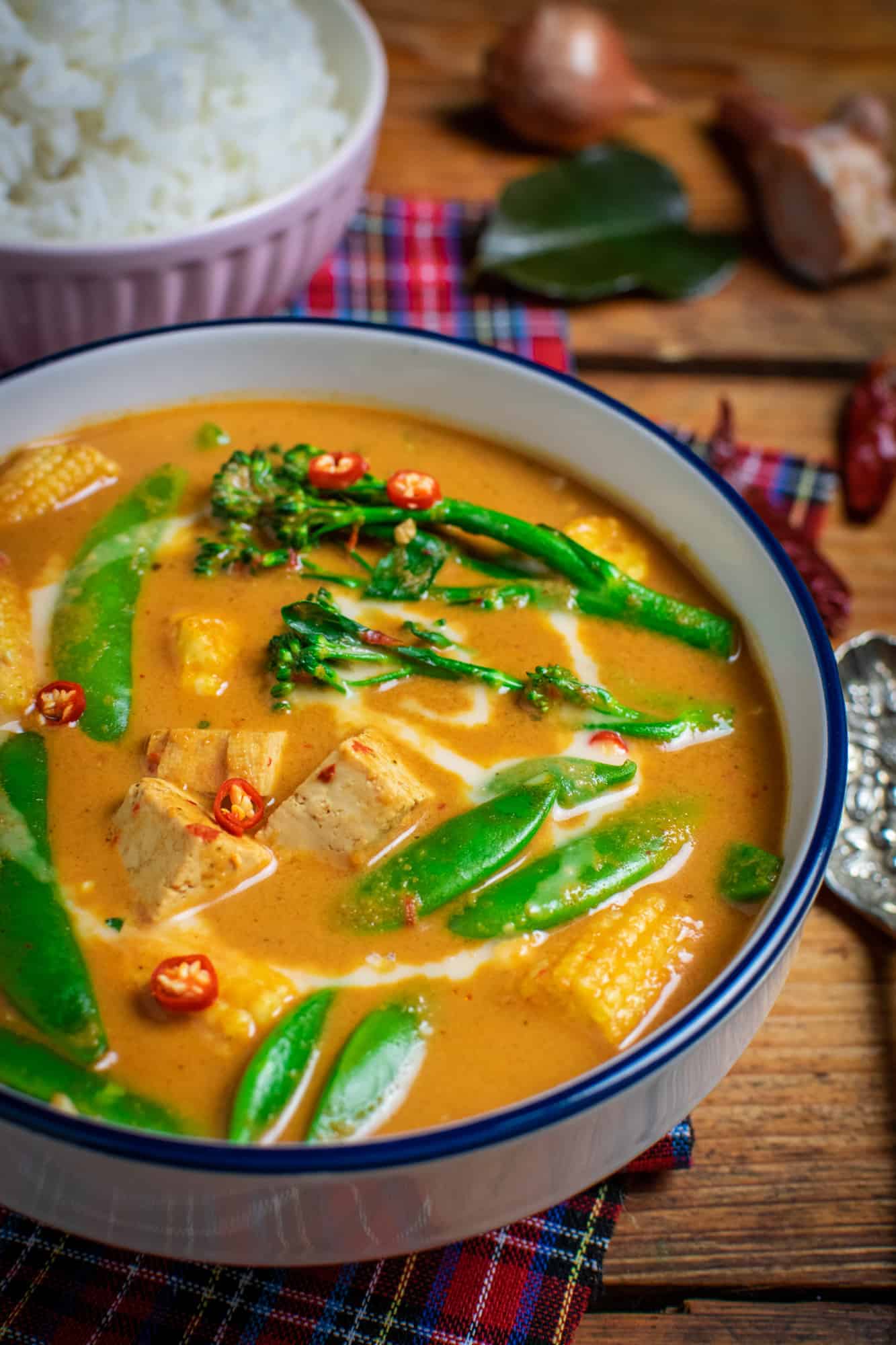 Thai red vegetable curry with rice in the background and ingredients such as Kaffir Lime leaves, chillies and onion