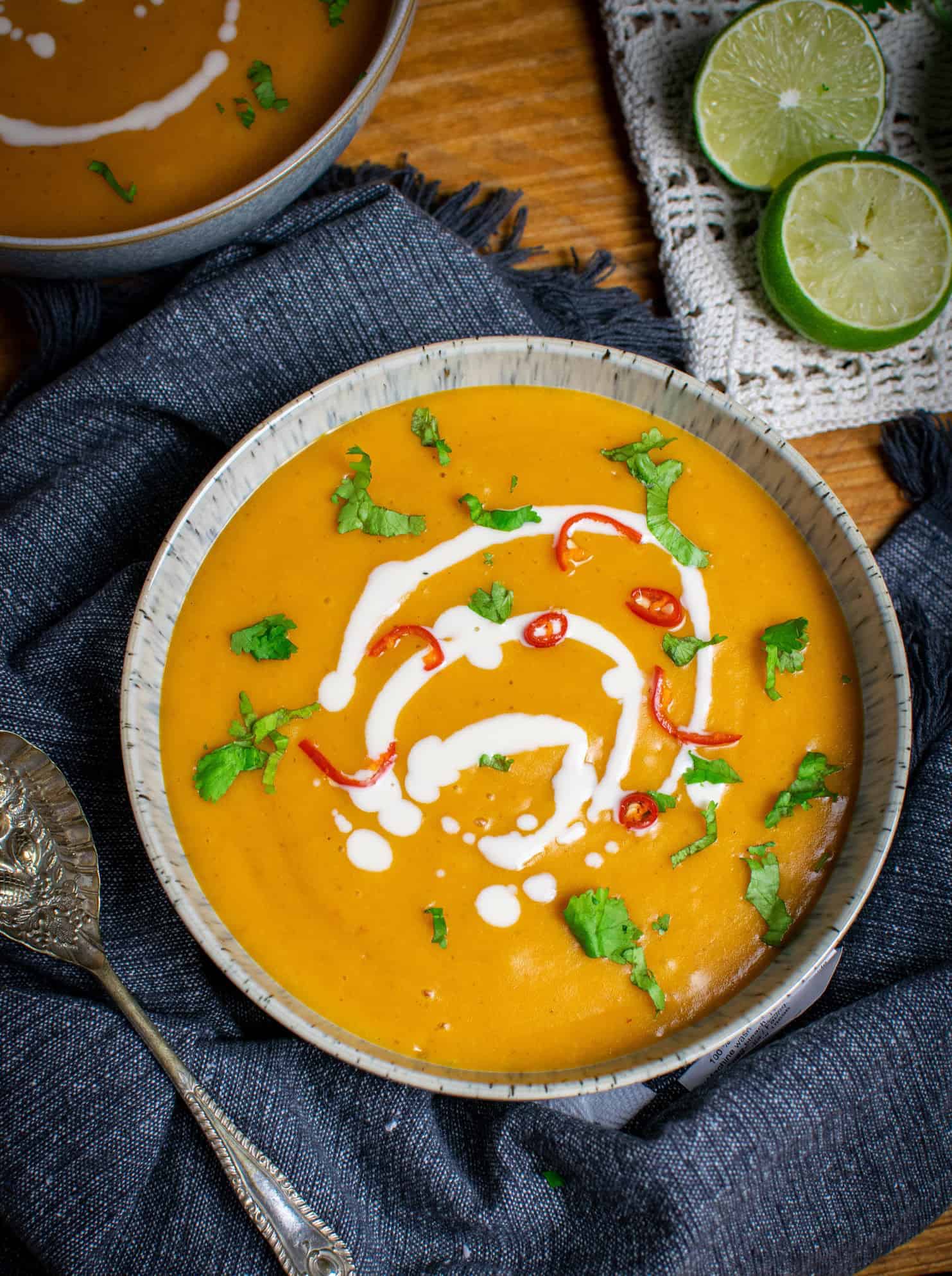 Vegan pumpkin soup with red curry paste, in a bowl on top of a blue towel, spoon to the left and lime in the background to the right