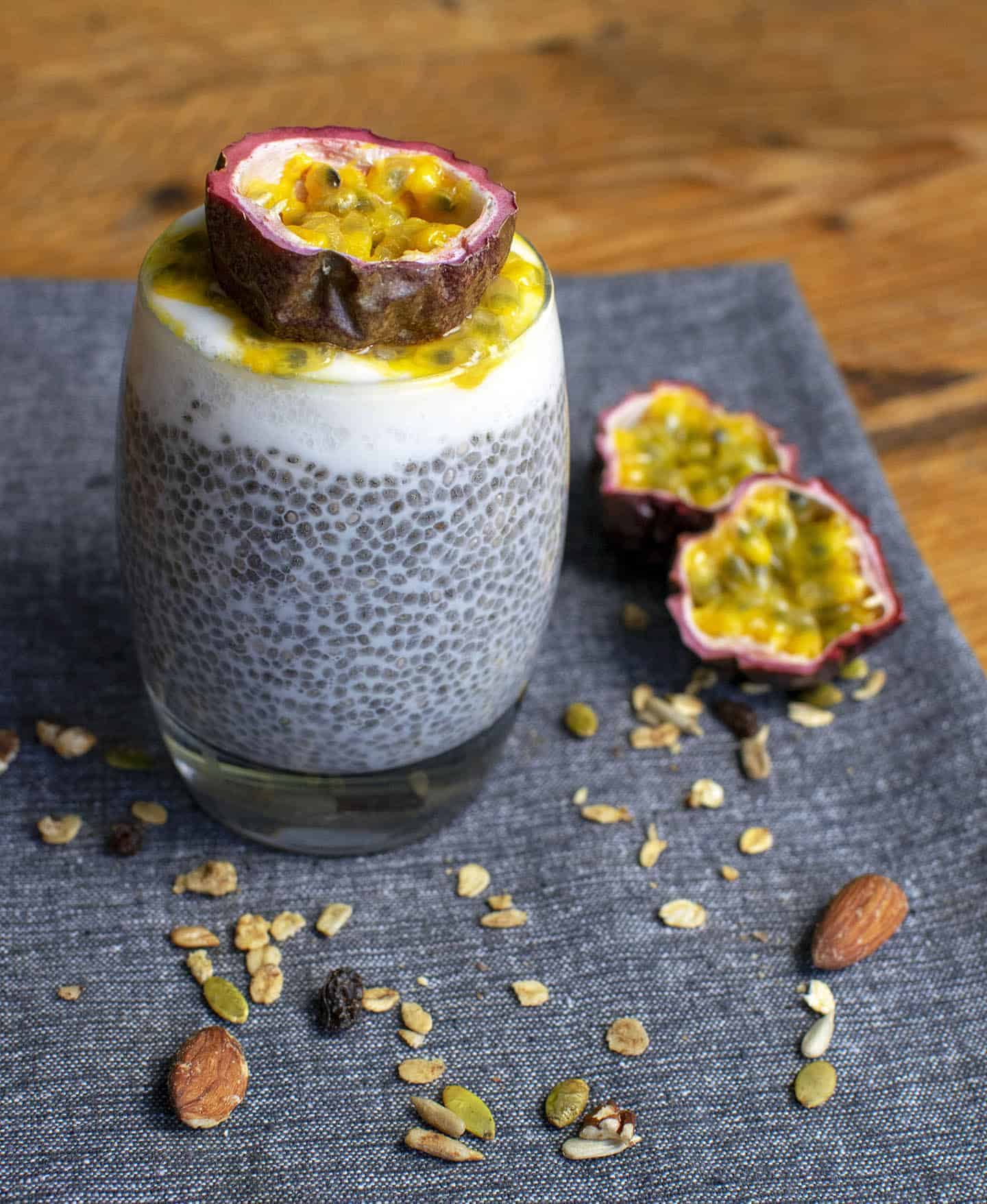 Passionfruit chia pudding set on a blue background in the second third of the portrait image. You can see the jelly-like chia seeds in the bulk of the glass with yoghurt and half a passionfruit on top. Granola is scattered around in the foreground.