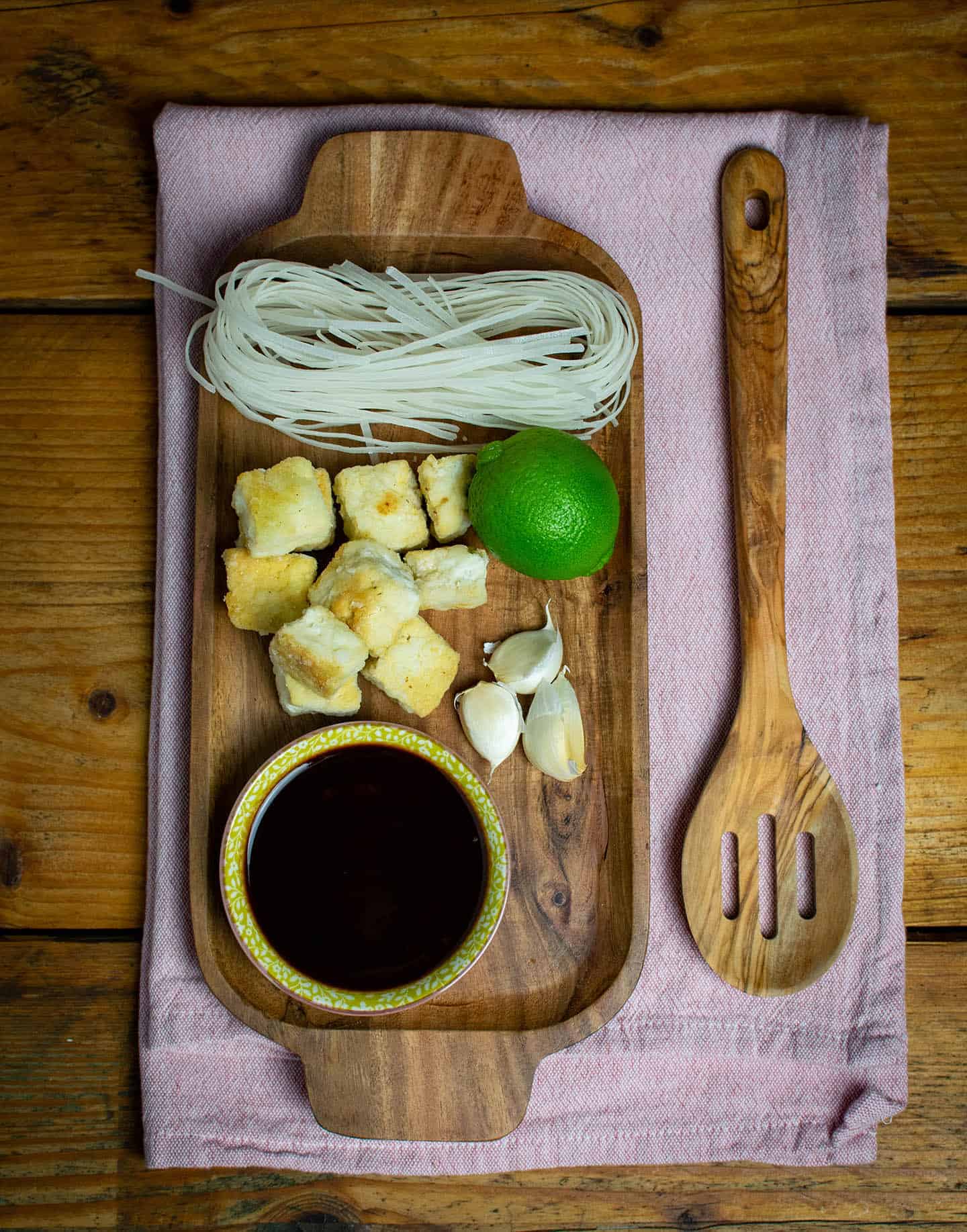 Ingredient laid out on a wooden board. Featuring noodles, tofu, lime and soy sauce.