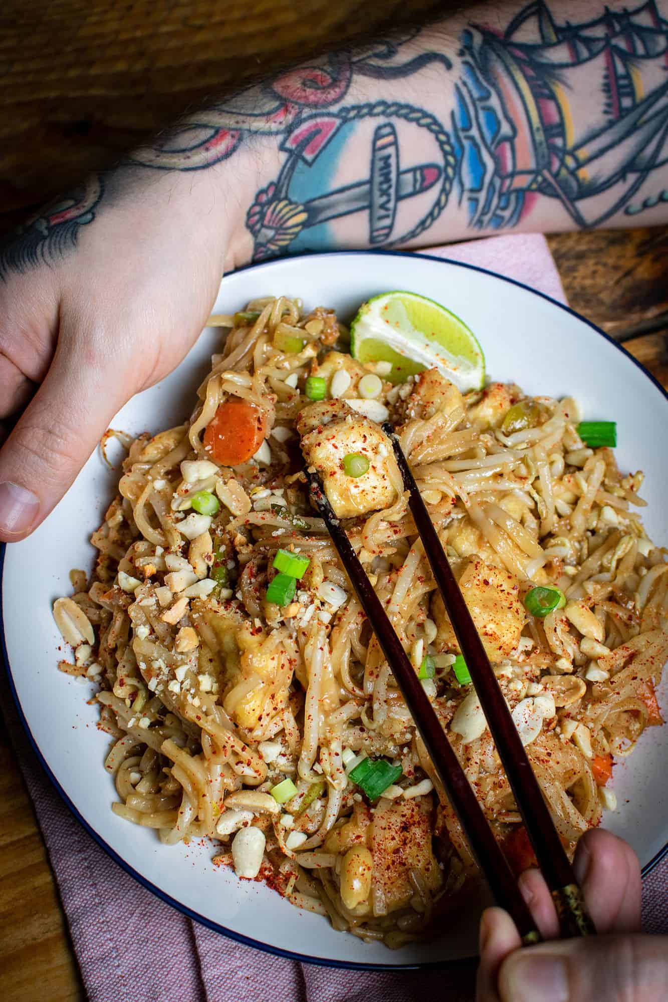 Easy vegan Pad Thai plated up with a tattooed arm holding the plate, and chopsticks holding a piece of tofu