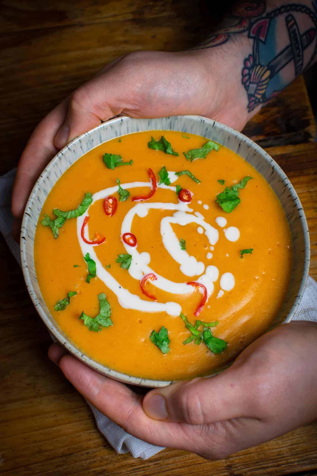 Vegan pumpkin soup in a bowl, held by two hands.Showing oat cream, fresh chilli and fresh coriander as toppings.