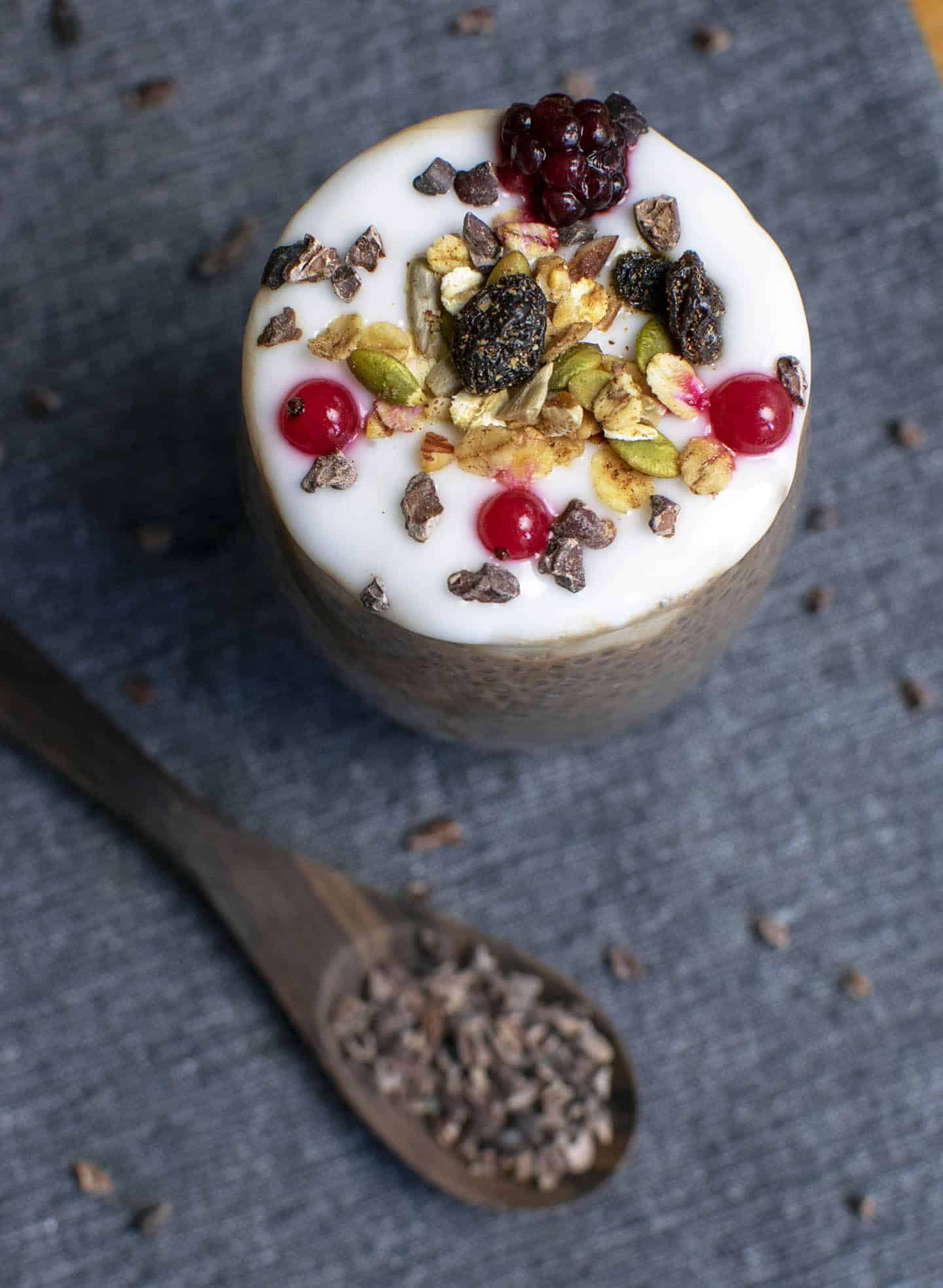 A scattering of vegan granola on top of a glass jar with red berries and blackcurrant. Set on a blue background with cacao nibs scattered on the background and a wooden spoon has cacao nibs placed on it.