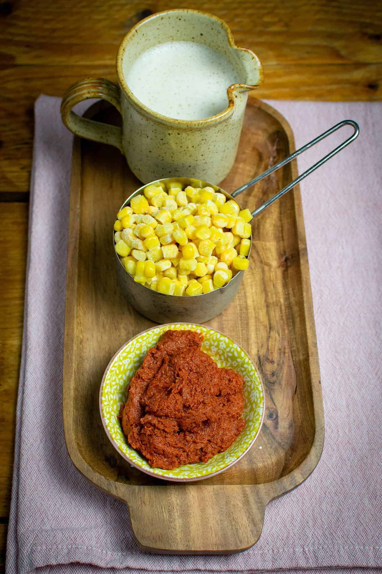 A slim wooden tray tray with a cream jar of oat milk, a cup measurer full with sweetcorn and Thai red curry paste in a small yellow dish.