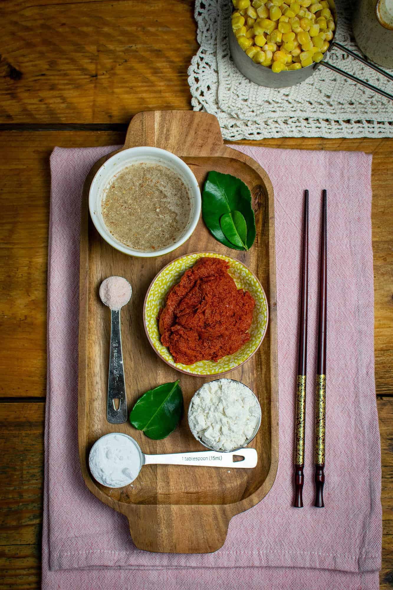 A slim wooden tray with lots of ingredients laid out on it, including baking powder, Thai curry paste, flour, kaffir lime leaves, salt, flax egg and sweetcorn in a cup measurer to in the right hand corner diagonal to the top of the tray.