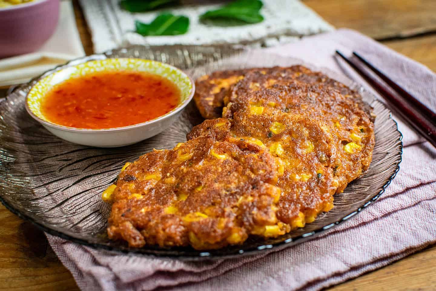 Close up of Thai sweetcorn fritters on a grey glass plate that's see through, Chopsticks to the right of the plate and sweet chilli sauce behind the fritters.