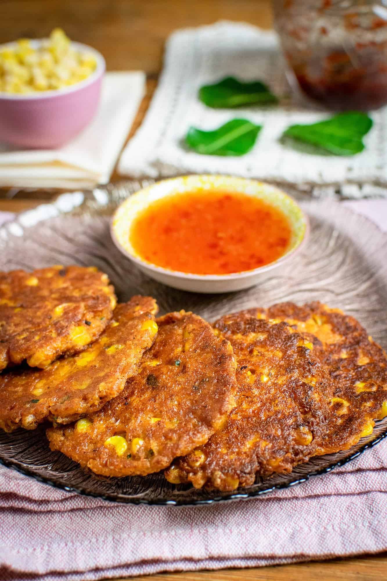 Thai sweetcorn fritters on a grey see-through plate with sweet chilli sauce in the background and a pot of sweetcorn and kaffir lime leaves for decoration.