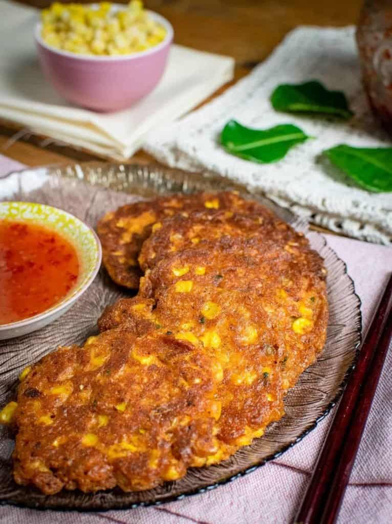 Grey glass plate with 5 corn fritters on it in an arc around a small yellow dish containing sweet chilli sauce. There's kaffir lime leaves in the background.