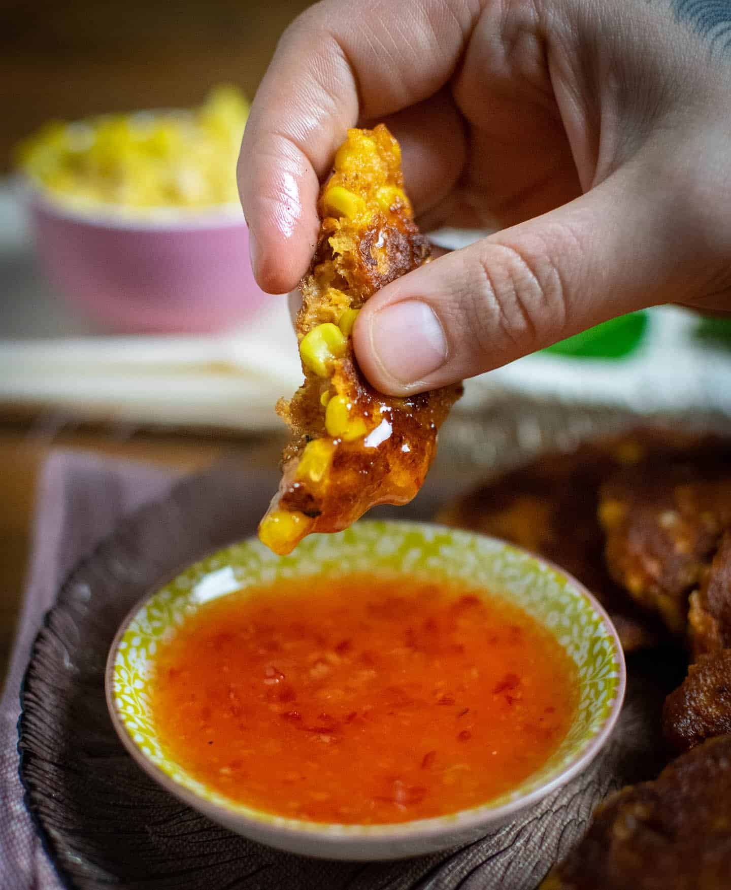 Dan's hand holding a piece of sweetcorn fritter after just dunking it in the pot of sweet chilli sauce beneath it. A pink pot full of sweetcorn is visible in the background.