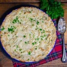 Landscape picture of a vegan fish pie in a circular dish, topped with mashed potato and parsley. There's a serving spoon to the right, a bunch of parsley in the background and the pie is resting on top of red checked material.