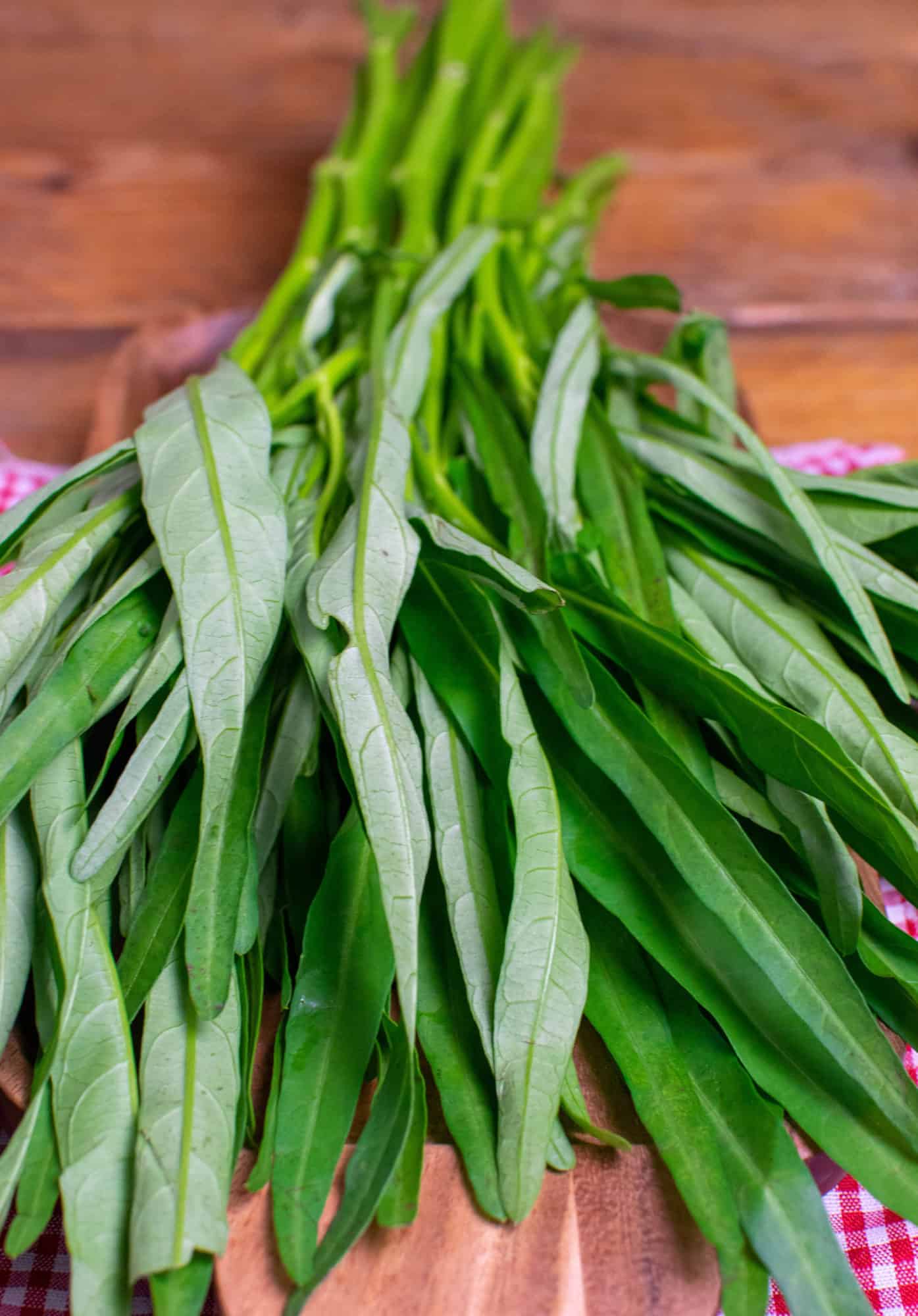 A fresh bunch of morning glory laying on a wooden board. The leafy end of the vegetable is closest to the camera, with the stalks running away in the background.