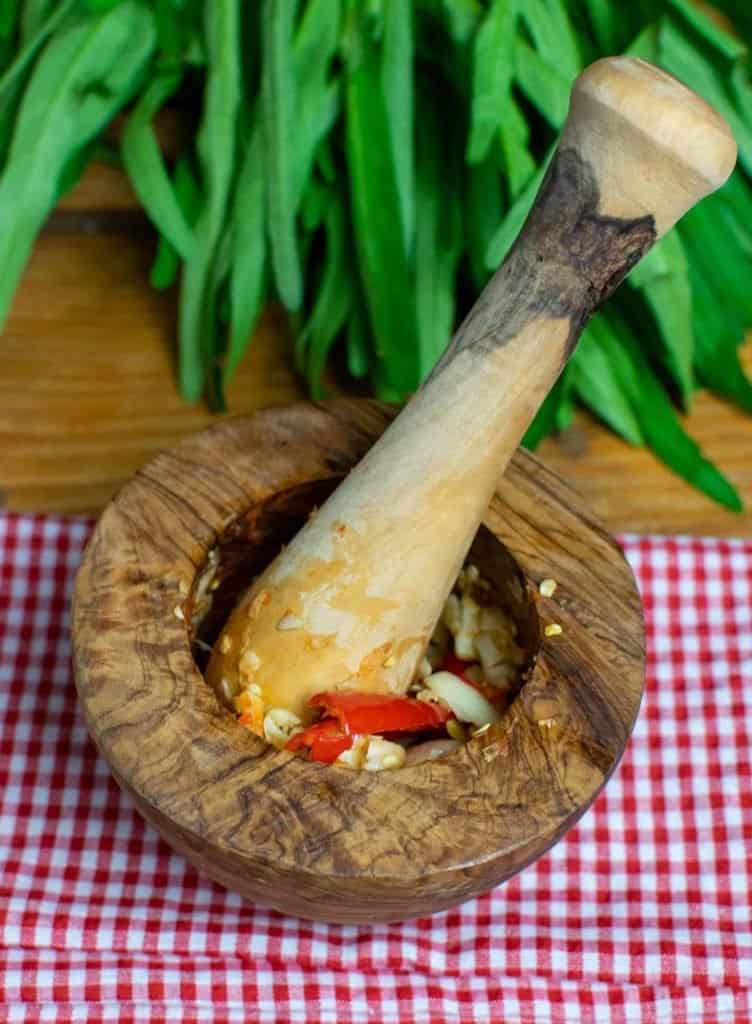 A wooden pestle and mortar sat on top of a gingham cloth. The mortar is filled with pounded garlic and red bird eye chillies. In the background of the image, at the top, is the leafy tops of a bunch of morning glory.