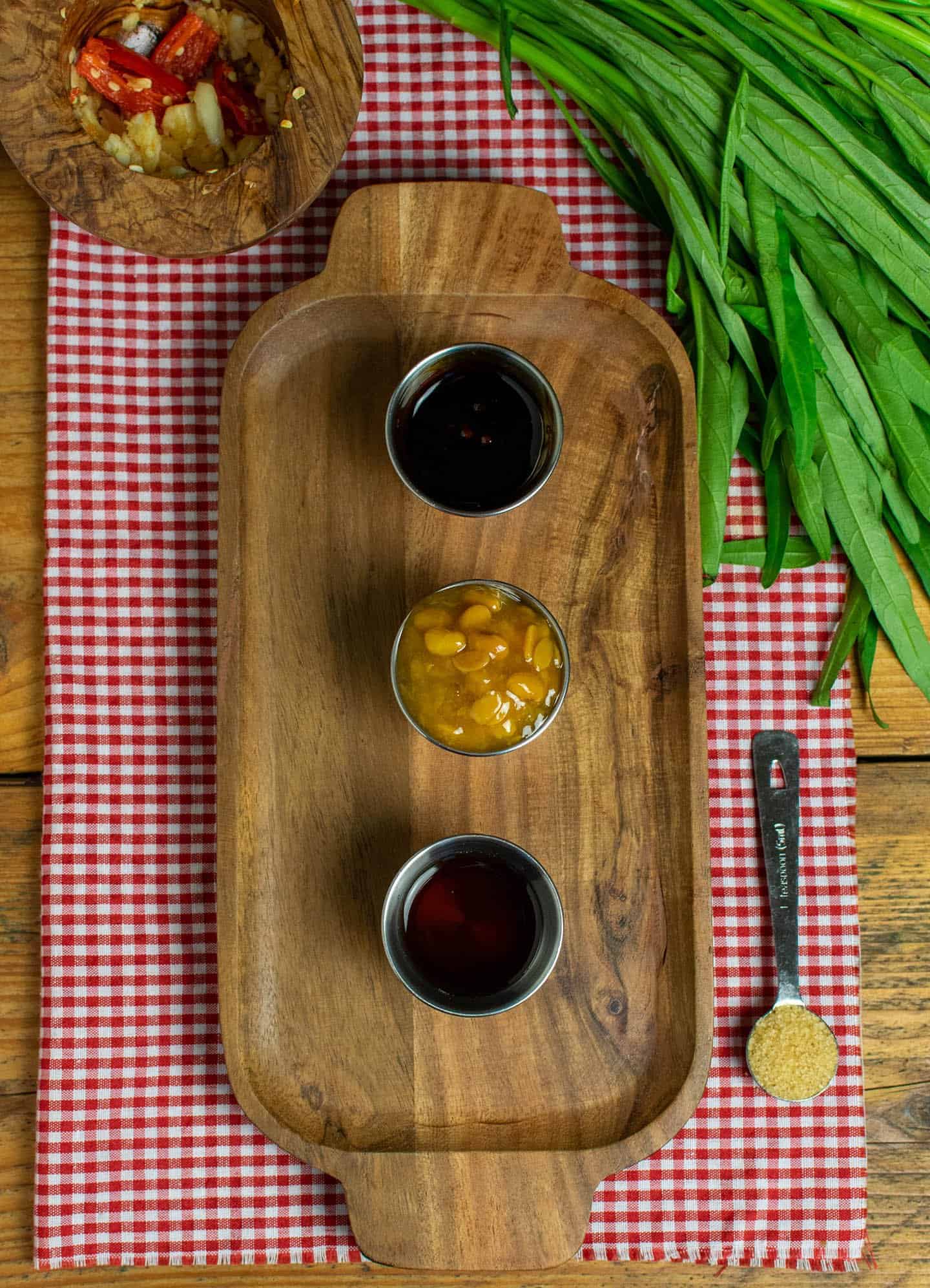 A photo of a wooden tray featuring some of the ingredients of Thai morning glory stir fry sits on top of a gingham cloth. Three pots are on the tray, filled with vegetarian oyster sauce, yellow bean sauce and vegetarian fish sauce. To sides of the tray are a tablespoon of sugar, a bunch of morning glory and a mortar filled with pounded garlic and chilli.