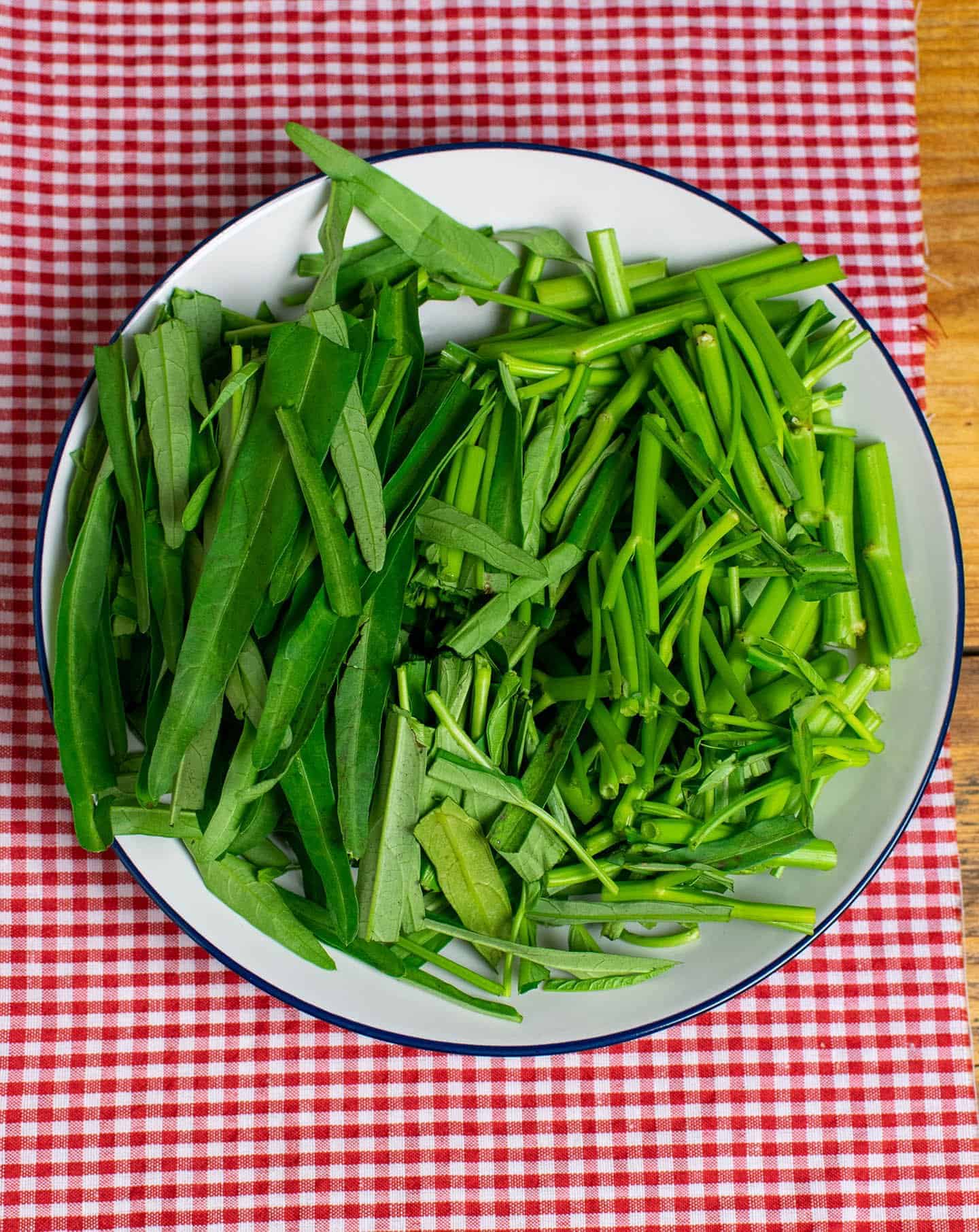 A white plate with a blue rim is filled with chopped morning glory. The pieces are 2-3 inches in length, with more stalks to the right of the plate and more of the leafy part of the vegetable to the left.