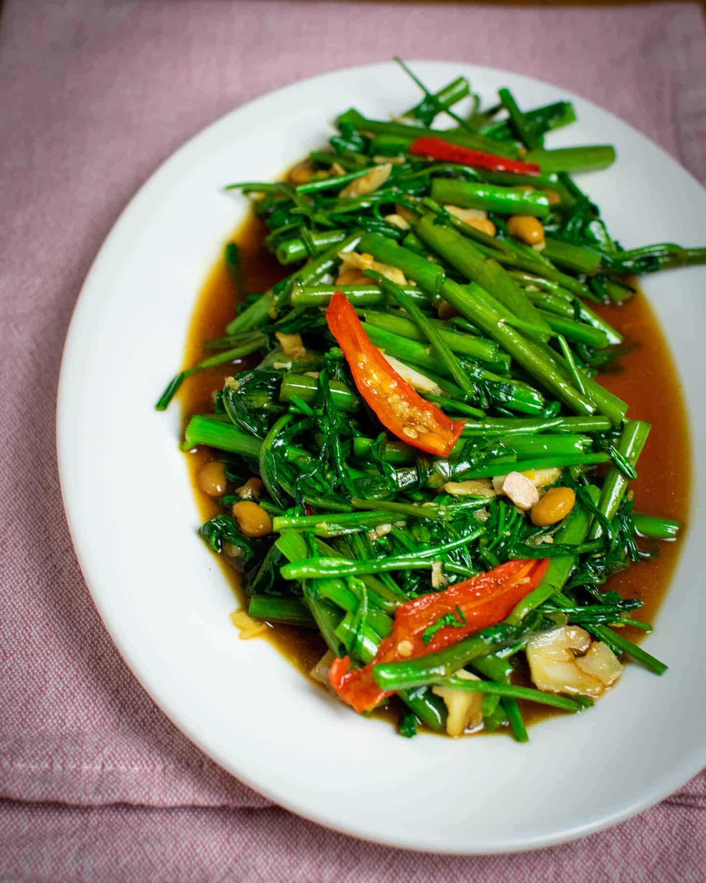 A white oval plate piled up with fresh Thai morning glory stir fry. The vibrant green stalks and leaves are piled high with pounded garlic and red chillies visible throughout, with the occasional soybean.