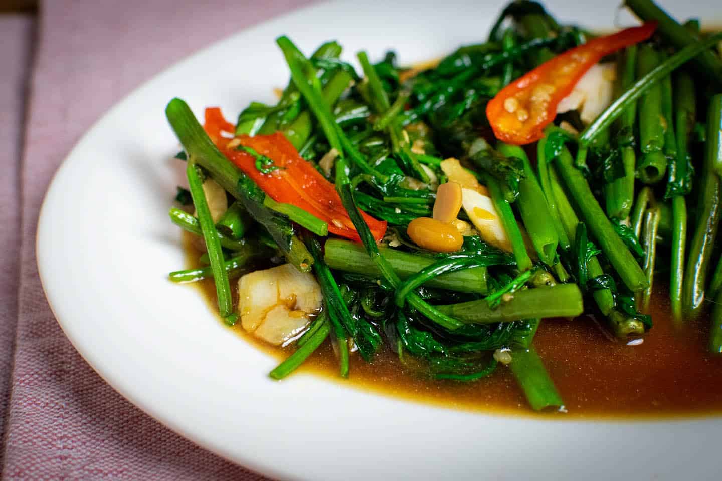 A close-up photo of stir fried morning glory. Several stalks and leaves are visible, with a couple of pieces of pounded up garlic, as well as sliced chillies and soybeans. A brown sauce sits underneath the vegetables and the edge of a white plate is visible underneath it all.