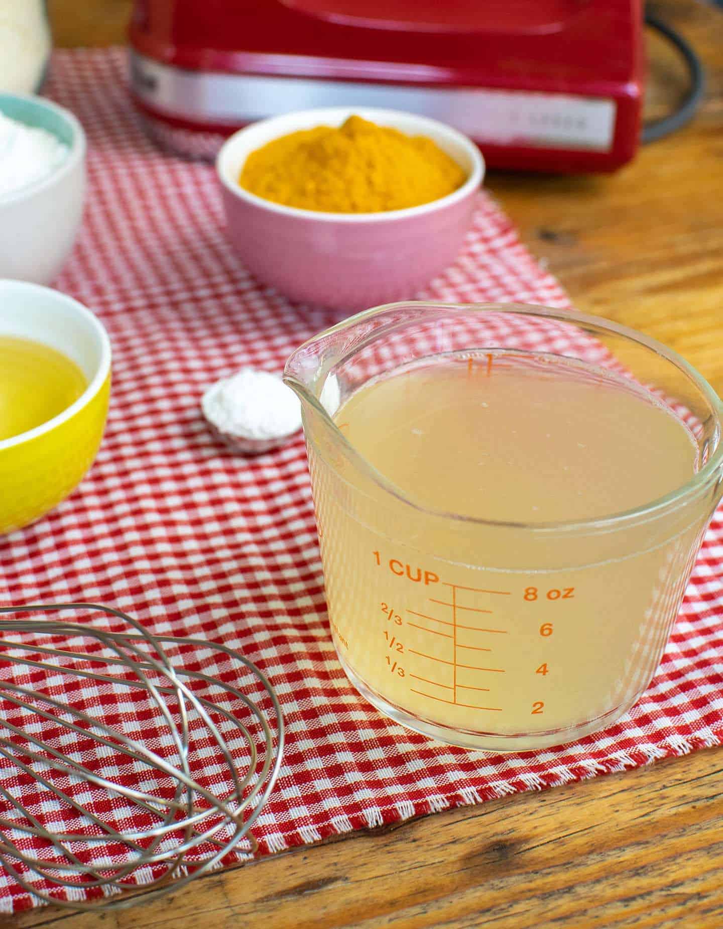 A photo of some of the ingredients that make vegan Yorkshire puddings. At the front is a measuring jug of aquafaba, with bowls of ground turmeric, vegetable oil and flour. There's also a tablespoon measurer filled with baking powder.