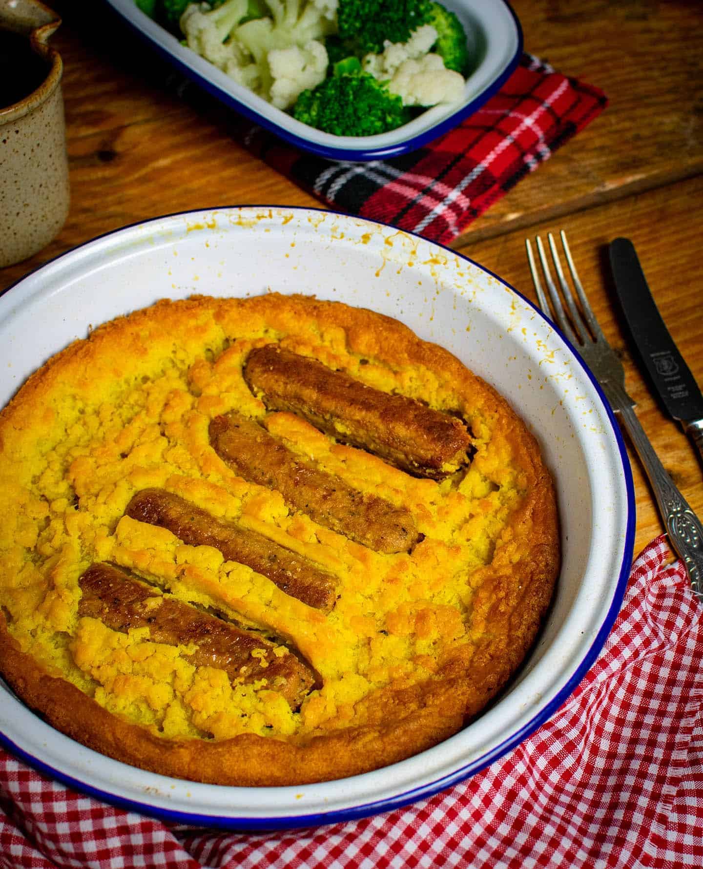 A photo of vegan toad in the hole in an enamel dish. A gingham cloth is wrapped round the dish and to the right hand side is a vintage knife and fork. Further back in the photo is an enamel side dish filled with broccoli and cauliflower, on top of a tartan cloth.
