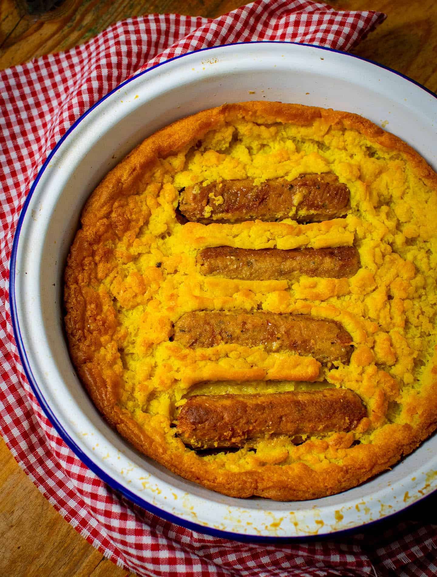 A top-down photo of vegan toad in the hole. It's in an enamel dish with a blue rim. Wrapped round the dish is a gingham cloth. The batter is gradients of yellow and golden brown, with 4 sausages poking through the surface.