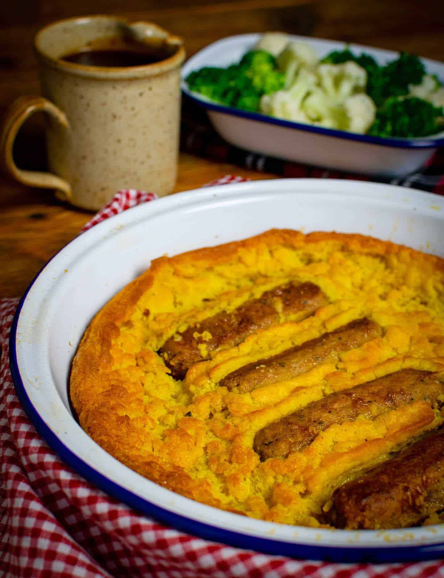 A baking pan filled with vegan toad in the hole. 4 sausages are visible and the edges of the batter are golden brown. In the background is a jug of gravy and a side dish of broccoli and cauliflower.