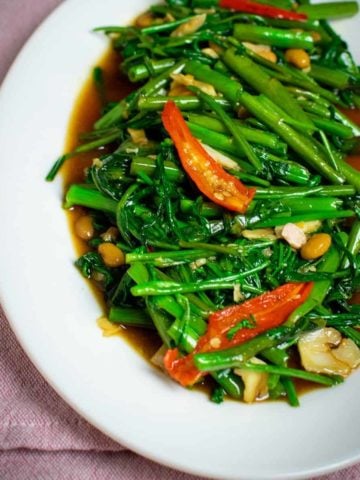 A large oval plate is filled with stir fried morning glory. The green shoots and leaves are spread across the plate, with the occasional piece of garlic, red chilli or soybean mixed in. Underneath the vegetables is a rich, brown sauce. The whole plate is sat on top of a pink cloth.