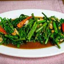 A landscape photo of a plate filled with stir fried morning glory. A rich brown sauce sits underneath a pile of fried morning glory, chunks of garlic, long red chillies and soybeans.