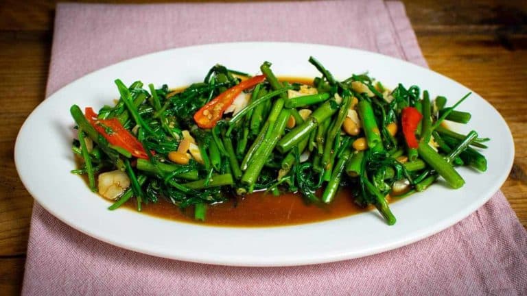A landscape photo of a plate filled with stir fried morning glory. A rich brown sauce sits underneath a pile of fried morning glory, chunks of garlic, long red chillies and soybeans.