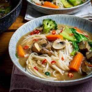 Noodles with gravy in a blue bowl with vegetables and red chilli on top. There is a second bowl in the background and a wok to the left in the background too.