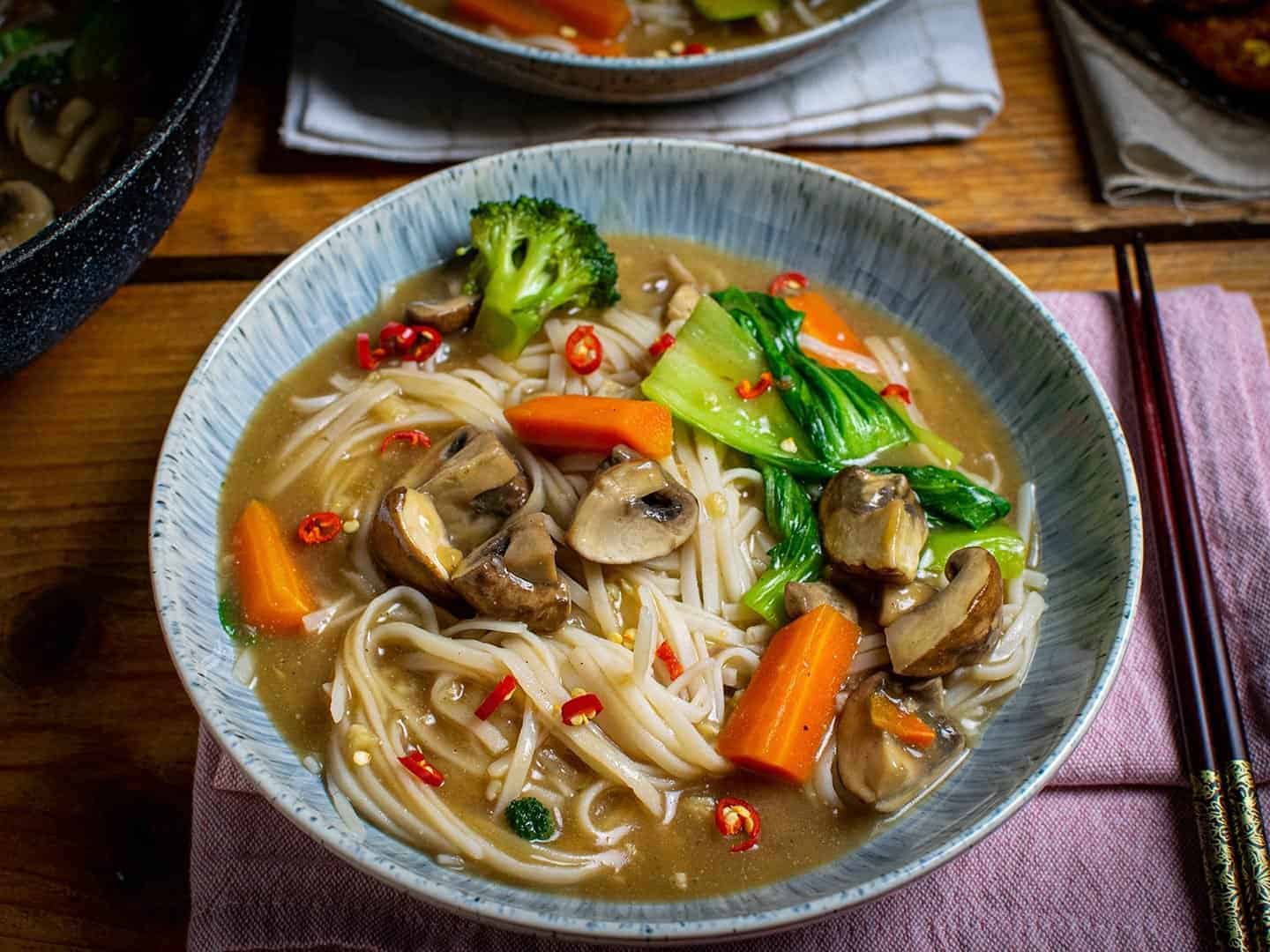 Noodles and veggies in the gravy sauce, served in a blue bowl with carrots, broccoli, pak choi and mushrooms are visible.