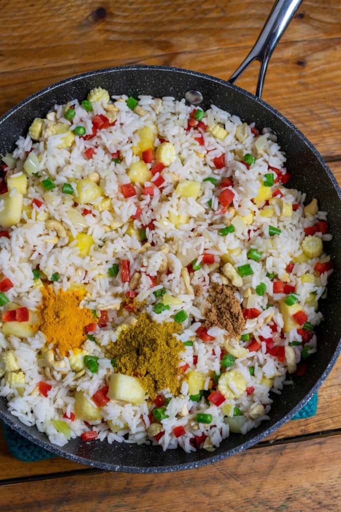 Un wok relleno de arroz, verduras en cubos y cúrcuma en polvo, curry en polvo y azúcar espolvoreado por encima en 3 pilas transparentes.